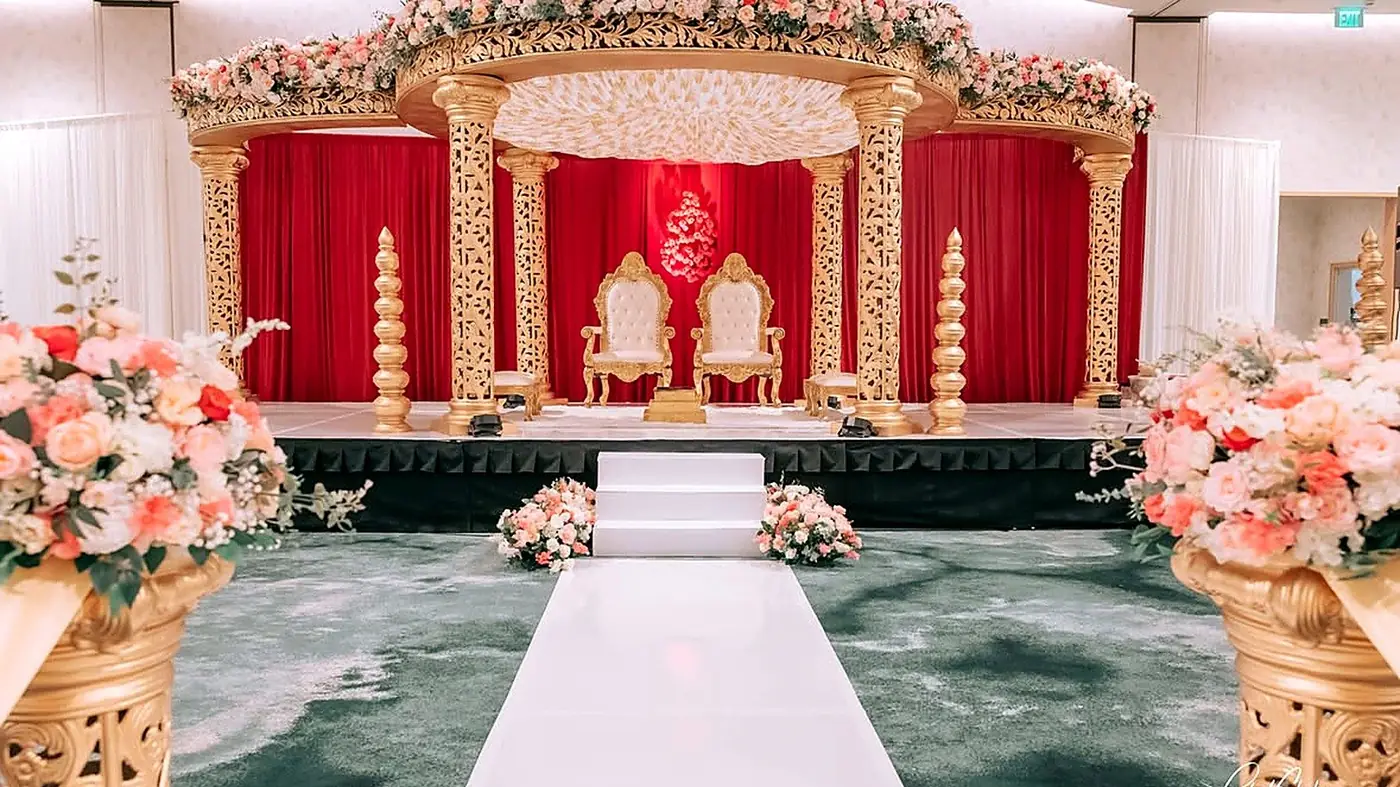 Gold carved mandap with red draping, floral canopy and twin thrones set on a skirted stage by Bsquare Events