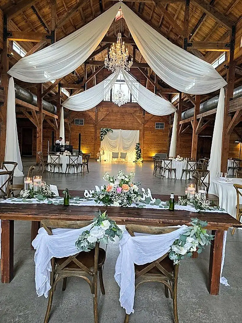 Barn wedding with white ceiling draping, crystal chandeliers and a florally styled sweetheart table by Bsquare Events