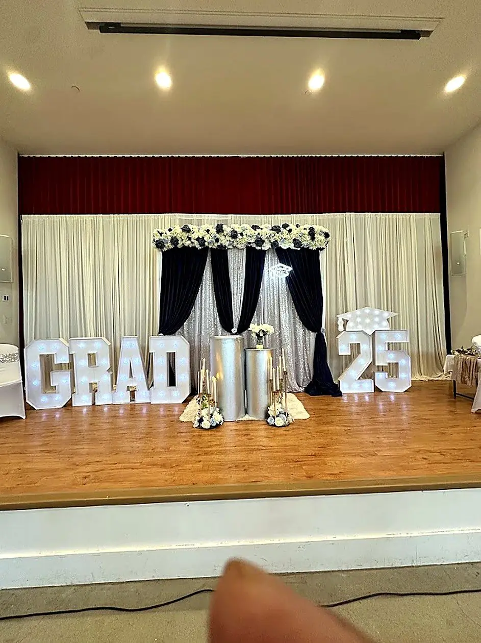 Graduation stage with navy and ivory draping, floral swag and illuminated GRAD 25 marquee letters by Bsquare Events