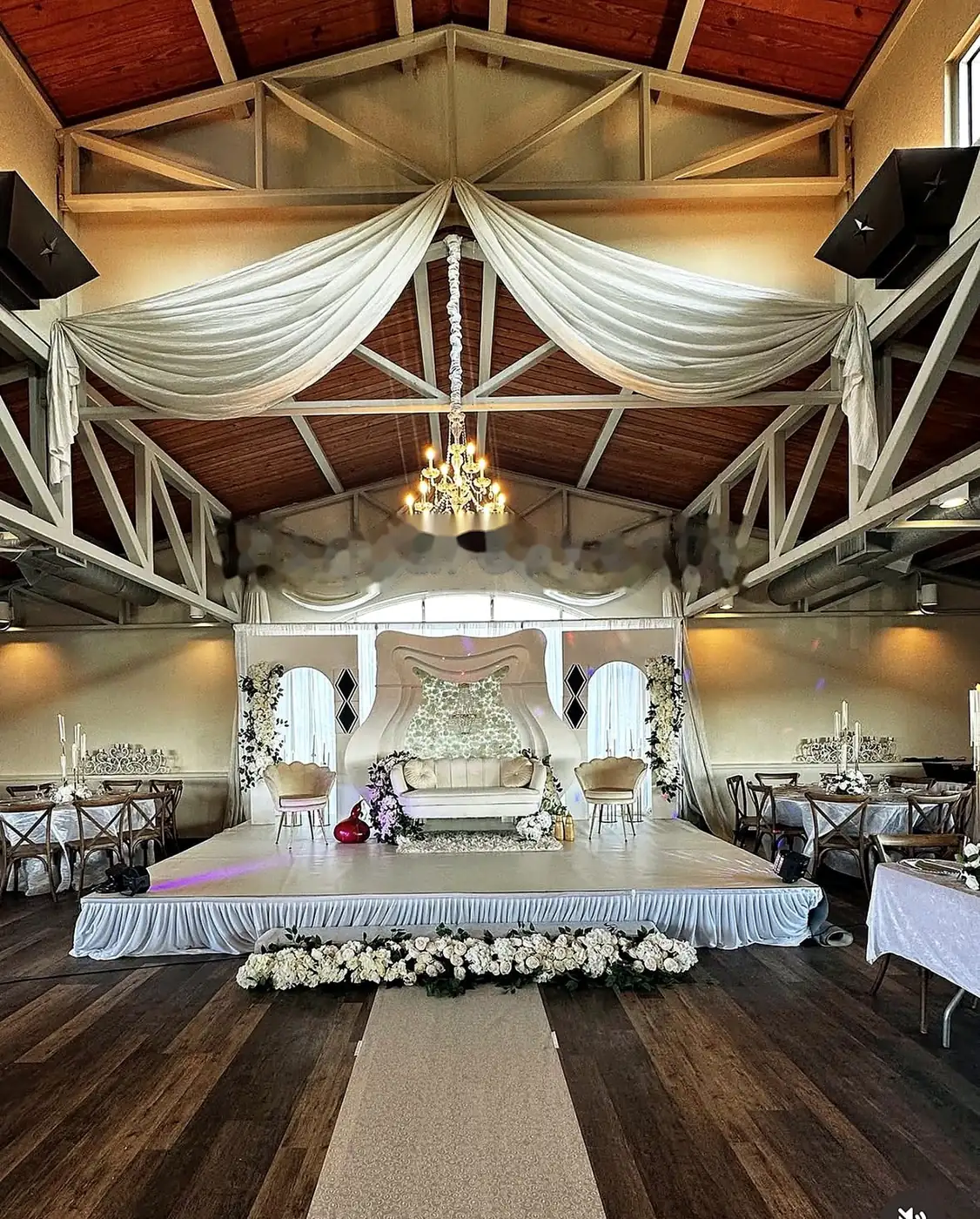 Barn wedding stage with sweeping ceiling draping, chandelier and a white floral reception backdrop by Bsquare Events