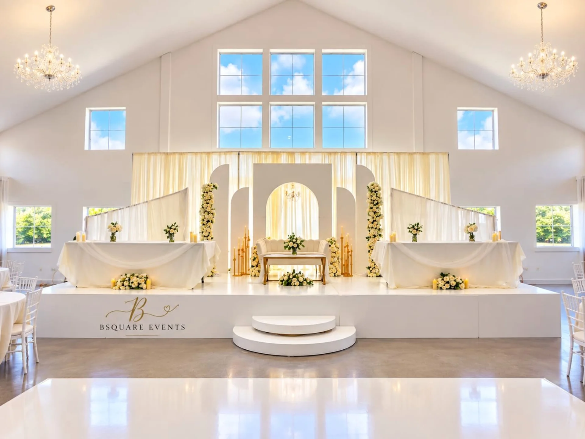 Elegant white and gold floral wedding stage with draped arches, chandeliers and the Bsquare Events logo in a bright ballroom by Bsquare Events